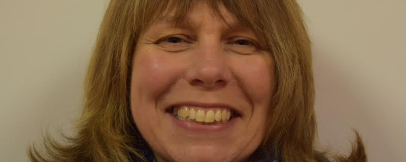 Smiling woman with shoulder-length hair wearing a blue scarf with a white pattern and a navy sweater against a plain background.