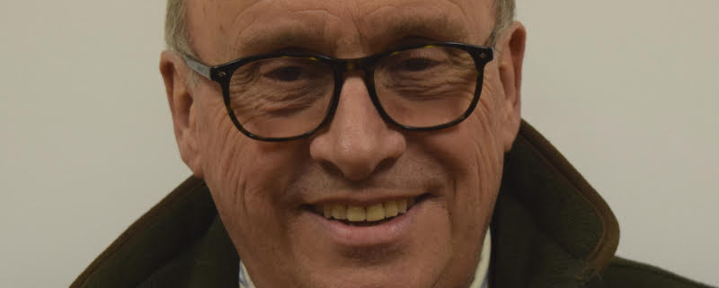 A man with glasses and a bald head is smiling. He is wearing a dark jacket over a collared shirt. The background is plain and light-colored.