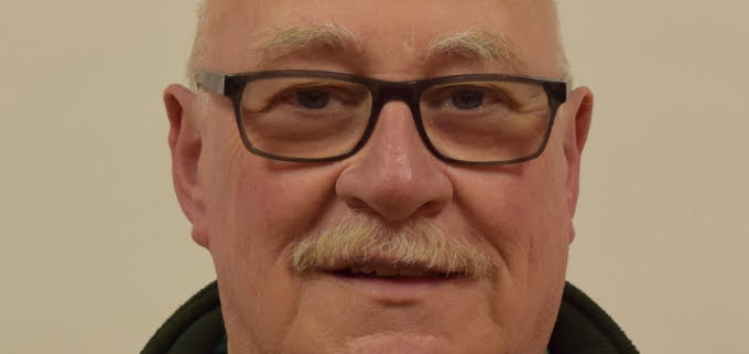 An older man with glasses, a mustache, and a bald head, wearing a dark green sweater and coat, smiles slightly against a plain background.
