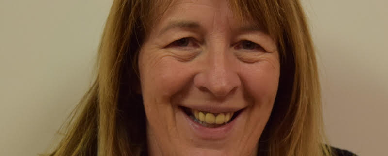 A woman with shoulder-length hair smiles at the camera. She is wearing glasses on her head, a blue jacket, and a light-colored shirt. The background is plain off-white.