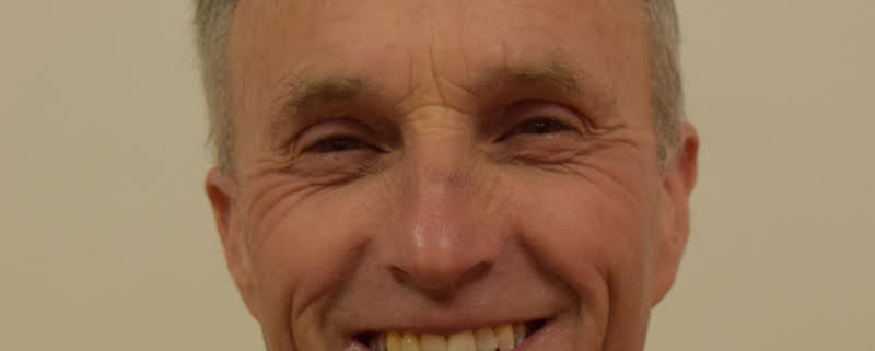 A smiling older man with thinning gray hair stands against a plain, beige background. He is wearing a dark, open-collared jacket and a black shirt. The man has a friendly expression with noticeable wrinkles around his eyes.