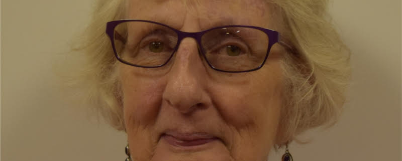 Elderly person with short white hair wearing glasses, a pink sweater, and dangling earrings against a plain background, smiling slightly.