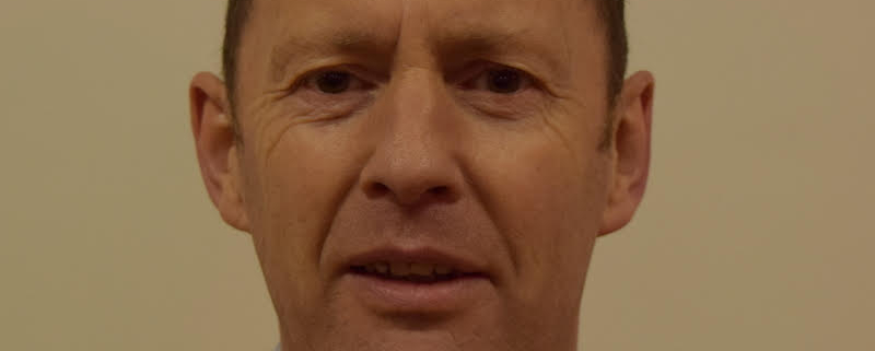 A man with short hair wearing a dark sweater over a collared shirt stands in front of a plain beige background, looking at the camera.