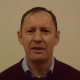 A man with short hair wearing a dark sweater over a collared shirt stands in front of a plain beige background, looking at the camera.