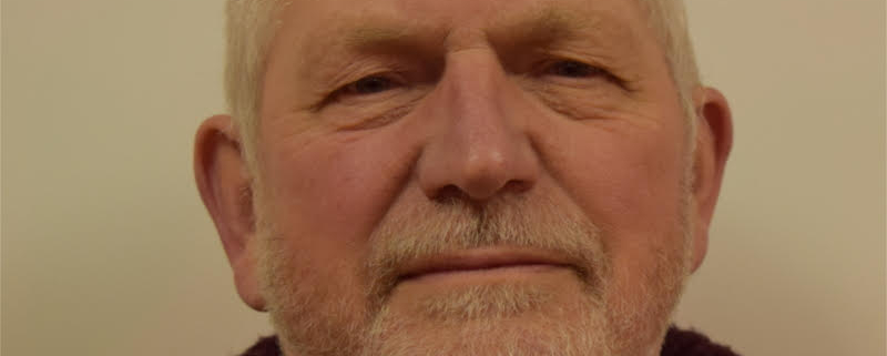 Portrait of an older man with a bald head, a short white beard, and a neutral expression. He is wearing a blue shirt and a brown sweater against a plain background.